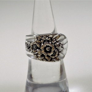 May include: A silver ring with a floral design. The ring is made from a repurposed spoon and features a large rose with smaller flowers surrounding it.