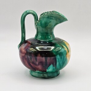 May include: A ceramic pitcher with a green handle and spout. The pitcher's body features a blend of colors, including green, purple, yellow, and black. The spout has a textured, scalloped edge. The pitcher sits on a small, round base.