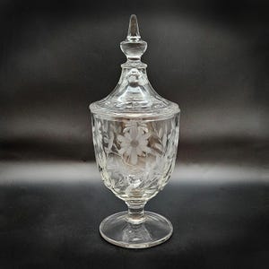 May include: A clear glass candy dish with a lid. The dish features etched floral designs, including daisies and leaves. It has a pedestal base and a pointed finial on the lid. The glass is transparent, allowing light to pass through.