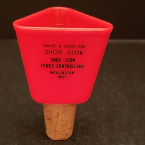 May include: A red plastic funnel-shaped bottle stopper with the text "HAVE A SHOT ON SHOX-STOK" and "SHOX-STOK FENCE CONTROLLERS WELLINGTON OHIO" printed in black. The stopper has a natural cork base.