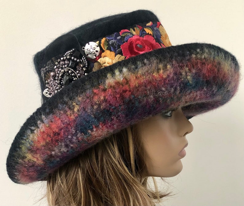 May include: A black felt hat with a wide brim and a colorful, embroidered band. The hat is decorated with a large, silver-toned brooch.