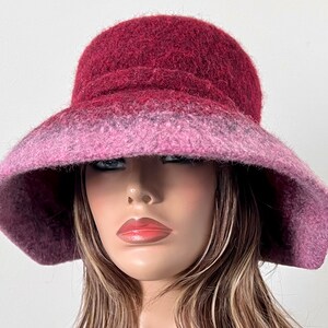 May include: A wide-brimmed hat made of felted wool in shades of burgundy and pink. The hat has a soft, fuzzy texture and a slightly slouched crown.