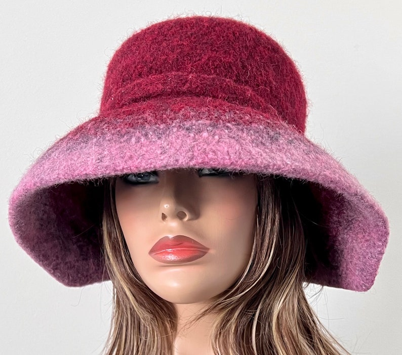 May include: A wide-brimmed hat made of felted wool in shades of burgundy and pink. The hat has a soft, fuzzy texture and a slightly slouched crown.