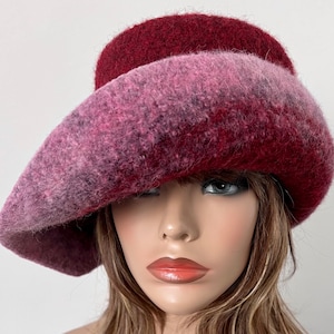 May include: A wide-brimmed hat made of felted wool in shades of pink and burgundy. The hat has a unique, asymmetrical shape and a soft, textured finish.