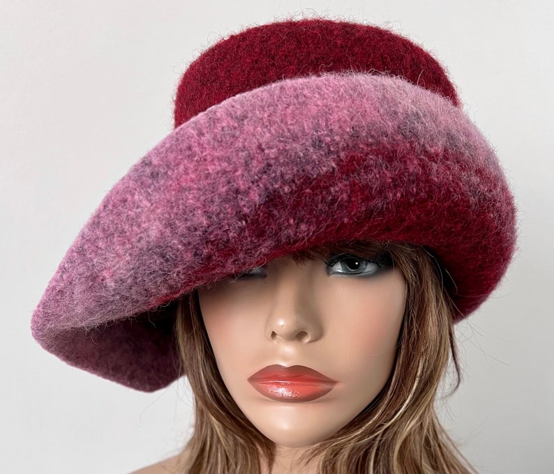 May include: A wide-brimmed hat made of felted wool in shades of pink and burgundy. The hat has a unique, asymmetrical shape and a soft, textured finish.