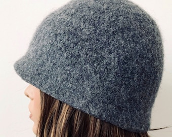 May include: A gray cloche hat with a small brim. The hat is made of a textured, wool-like material. The hat is shown from a side profile.