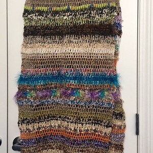 May include: A multi-colored crocheted scarf with fringe. The scarf has stripes of pink, green, brown, beige, blue, purple, orange, and gold. The scarf is hanging on a white door.