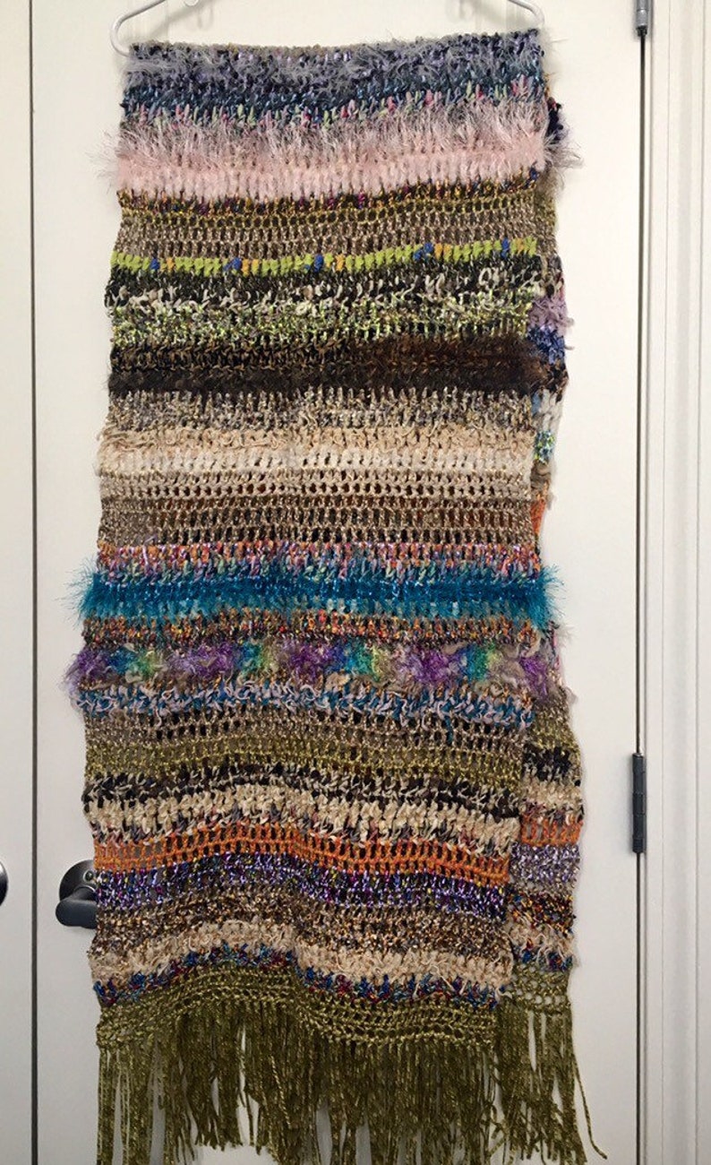 May include: A multi-colored crocheted scarf with fringe. The scarf has stripes of pink, green, brown, beige, blue, purple, orange, and gold. The scarf is hanging on a white door.