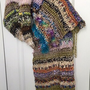 May include: A multi-colored crocheted shawl with fringe. The shawl is made with a variety of yarns, including some with a fuzzy texture. The shawl is a mix of brown, green, orange, purple, blue, and white.