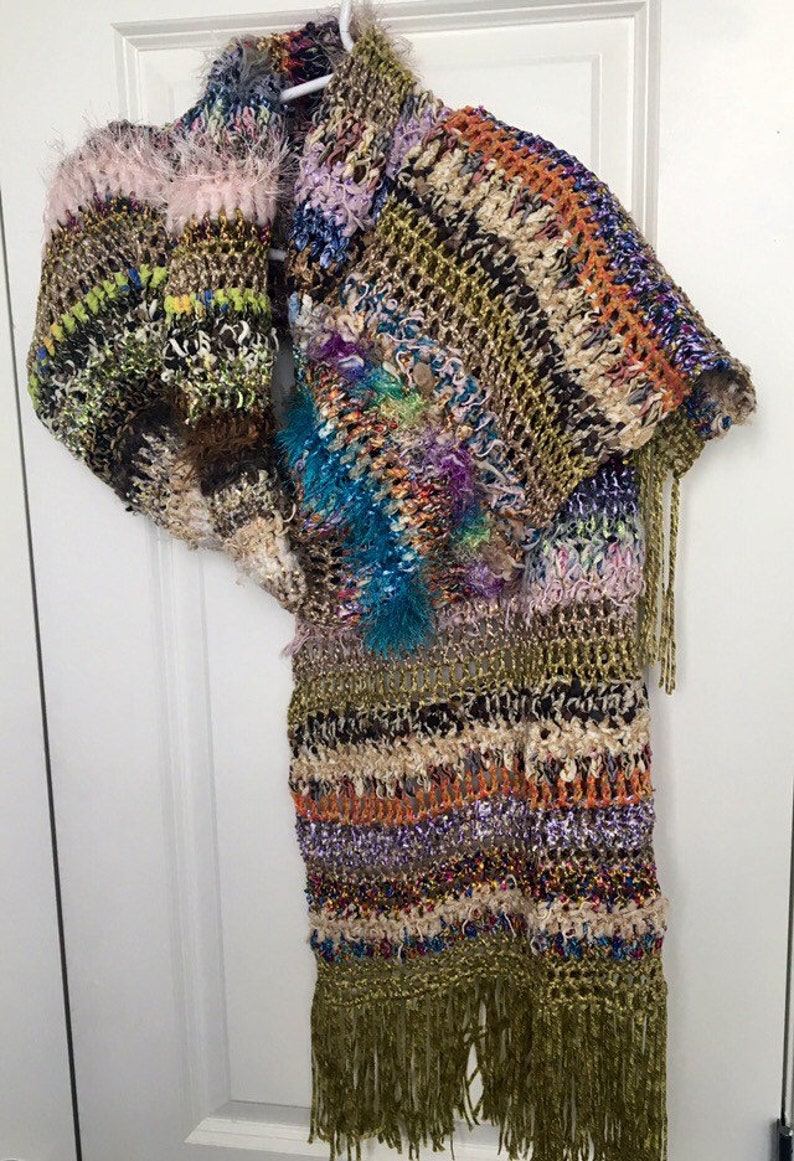 May include: A multi-colored crocheted shawl with fringe. The shawl is made with a variety of yarns, including some with a fuzzy texture. The shawl is a mix of brown, green, orange, purple, blue, and white.