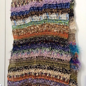 May include: A multi-colored crocheted scarf with fringe. The scarf is made with a variety of yarns, including fuzzy, sparkly, and textured yarns. The scarf is a mix of brown, green, blue, purple, orange, and white.