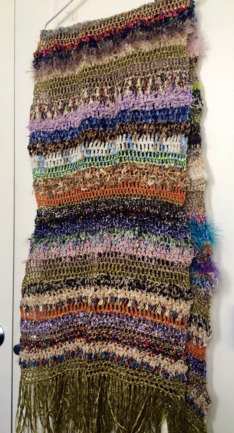 May include: A multi-colored crocheted scarf with fringe. The scarf is made with a variety of yarns, including fuzzy, sparkly, and textured yarns. The scarf is a mix of brown, green, blue, purple, orange, and white.