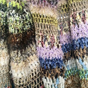 May include: Close-up of a multi-colored crocheted fabric with a variety of textures, including fuzzy, looped, and woven. The fabric is made up of different shades of brown, green, purple, blue, and white.