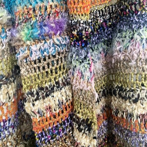 May include: Close-up of a multi-colored crocheted fabric with a variety of textures and patterns. The fabric is made with a mix of yarns, including gold, orange, blue, and white.