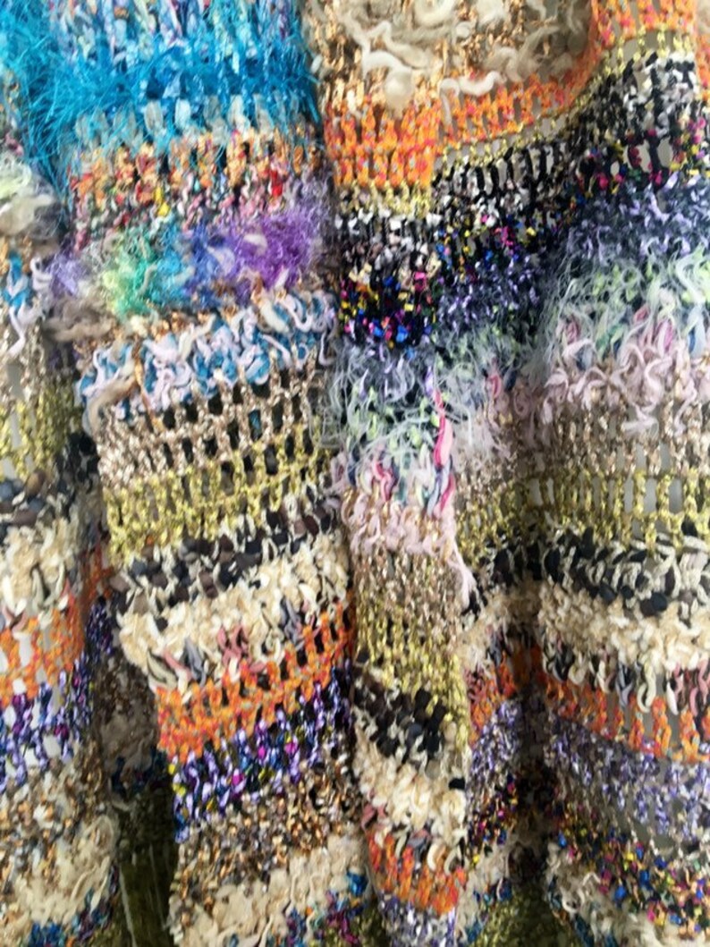 May include: Close-up of a multi-colored crocheted fabric with a variety of textures and patterns. The fabric is made with a mix of yarns, including gold, orange, blue, and white.
