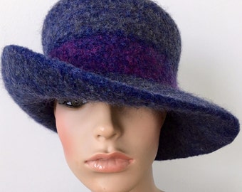 Puede incluir: Un sombrero de ala ancha hecho de lana felpuda en tonos azules y morados. El sombrero tiene un acabado suave y texturizado y un ala ancha que se puede peinar de varias maneras.