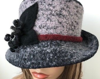 May include: A black and gray felt hat with a burgundy band and a black felt flower accent. The hat has a wide brim and a slightly rounded crown.