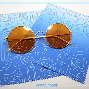 Puede incluir: Gafas de sol redondas de color naranja con montura de metal plateado sobre un pa&ntilde;o de microfibra de limpieza con estampado azul y blanco. El pa&ntilde;o tiene un borde blanco y un borde festoneado.