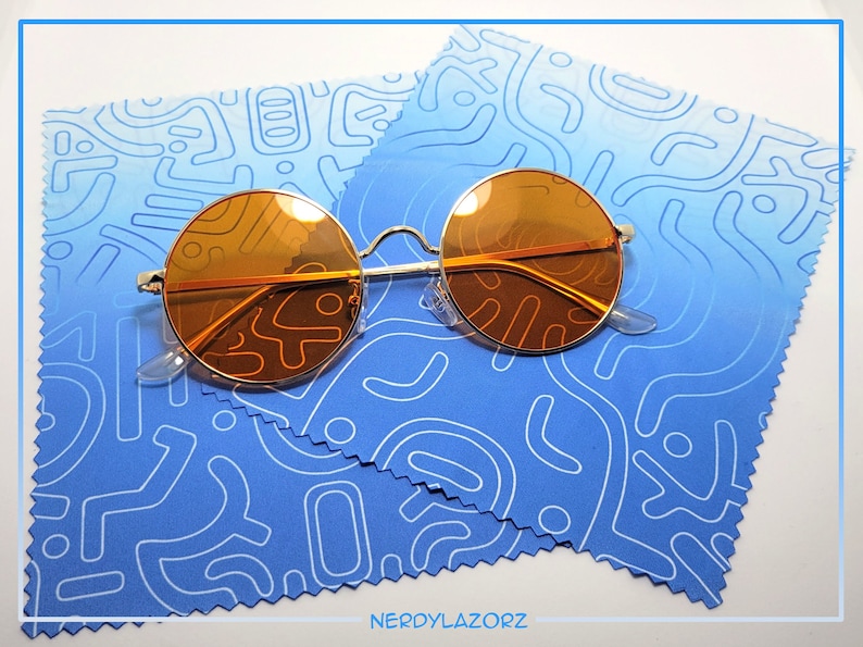 Puede incluir: Gafas de sol redondas de color naranja con montura de metal plateado sobre un pa&ntilde;o de microfibra de limpieza con estampado azul y blanco. El pa&ntilde;o tiene un borde blanco y un borde festoneado.