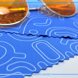 Puede incluir: Un par de gafas de sol redondas de color naranja sobre un pa&ntilde;o de microfibra azul con un patr&oacute;n abstracto blanco. El pa&ntilde;o tiene un borde en zigzag.