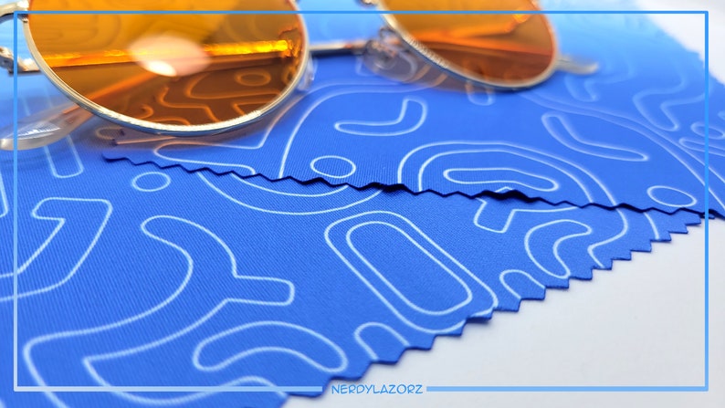 Puede incluir: Un par de gafas de sol redondas de color naranja sobre un pa&ntilde;o de microfibra azul con un patr&oacute;n abstracto blanco. El pa&ntilde;o tiene un borde en zigzag.