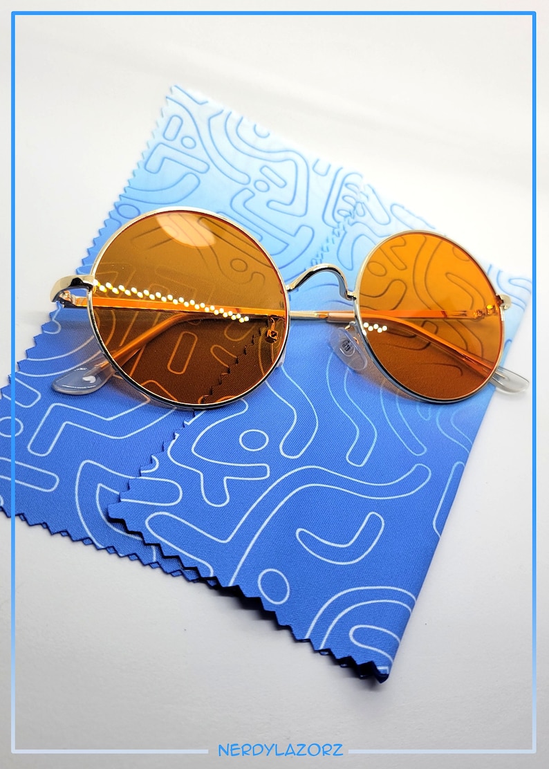Puede incluir: Gafas de sol redondas de color naranja con montura de metal dorado, apoyadas sobre un pa&ntilde;o de microfibra azul con un patr&oacute;n abstracto blanco. El pa&ntilde;o tiene un borde festoneado.
