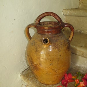 May include: A large, brown earthenware jug with two handles and a spout. The jug has a glazed finish and is sitting on a set of stone steps.