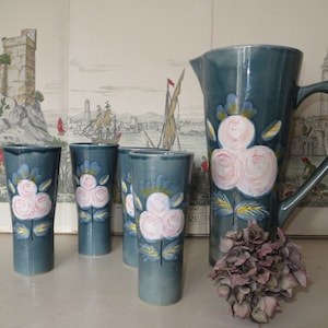 May include: Set of four blue ceramic drinking glasses with white and pink rose designs and a matching blue ceramic pitcher with a white and pink rose design.