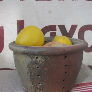 May include: A round, brown, rustic ceramic colander with small drainage holes along the sides and a thick rim. The bowl contains two bright yellow lemons and a light brown onion. The exterior surface has a weathered, textured appearance.