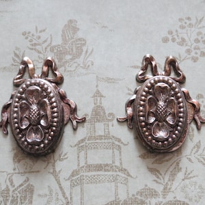May include: Two identical oval-shaped metallic ornaments with a copper-toned finish. Each piece features a central embossed floral motif surrounded by a beaded border, topped with a decorative bow element with flowing ribbons extending to the sides.