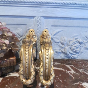 Peut inclure: Deux consoles décoratives dorées avec un motif ornemental en forme de volute. Elles sont placées contre un mur bleu clair avec des sculptures florales en relief et reposent sur une surface en marbre.