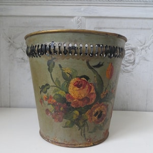 Könnte beinhalten: Ein Abfalleimer im Vintage-Stil mit einem verwitterten, salbeigrünen Finish. Der Metalleimer hat einen goldenen Rand und ein dekoratives Blumendesign mit Rosen in Gelb-, Orange- und Rottönen. Der obere Rand hat eine Reihe kleiner, gleichmäßig verteilter Löcher.