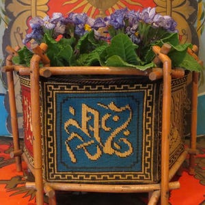 May include: A decorative planter with a tapestry design, featuring a blue and gold central motif. The planter is supported by a wooden frame and filled with purple flowers and green foliage. The design is ornate and decorative, suitable for a home.