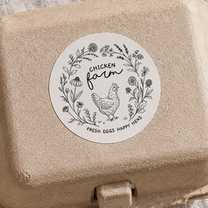 May include: A cardboard egg box with a circular "Chicken Farm" sticker, next to an open egg box with several eggs. The sticker features a hen illustration and the text "Fresh Eggs Happy Hens". The eggs and box are light brown.