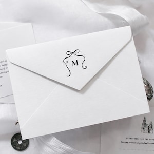 May include: A white envelope featuring a black bow and the letter "M". The envelope is part of a wedding invitation set, with other cards visible in the background, suggesting a formal event.