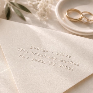 May include: Ivory-colored wedding invitation envelopes with embossed text reading "George + Olive, 1703 Broadway Market, New York, NY 98001." A pair of gold wedding rings rests on a small white dish, with floral accents.