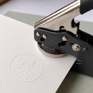 K&ouml;nnte beinhalten: Ein manuelles Pr&auml;gewerkzeug aus schwarzem und silbernem Metall, das ein kreisf&ouml;rmiges Motiv auf ein wei&szlig;es Blatt Papier dr&uuml;ckt. Der Pr&auml;geabdruck zeigt den Text Your logo here in einer Schreibschrift innerhalb eines d&uuml;nnen Kreisrandes.