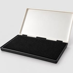 Könnte beinhalten: Ein rechteckiges Stempelkissen mit einer schwarzen Filzoberfläche in einem schwarzen Kunststoffgehäuse. Der befestigte Deckel ist innen weiß und verfügt über ein Scharnierdesign, wodurch sich das Gehäuse vollständig öffnen lässt.