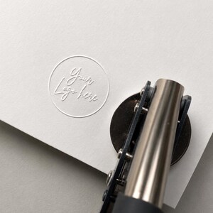 K&ouml;nnte beinhalten: Ein manuelles Pr&auml;gewerkzeug aus Metall, das auf ein dickes wei&szlig;es Blatt Papier dr&uuml;ckt. Das Papier zeigt ein kreisf&ouml;rmiges Pr&auml;gemotiv mit dem Text Your Logo here in Schreibschrift. Das Werkzeug hat einen dunklen Griff und eine silberne Oberfl&auml;che.