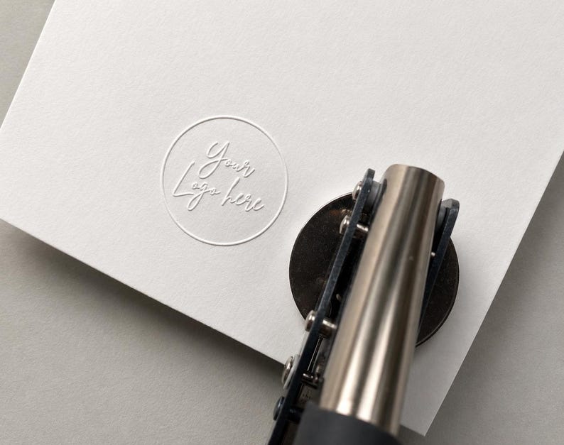 K&ouml;nnte beinhalten: Ein manuelles Pr&auml;gewerkzeug aus Metall, das auf ein dickes wei&szlig;es Blatt Papier dr&uuml;ckt. Das Papier zeigt ein kreisf&ouml;rmiges Pr&auml;gemotiv mit dem Text Your Logo here in Schreibschrift. Das Werkzeug hat einen dunklen Griff und eine silberne Oberfl&auml;che.