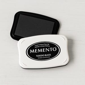Könnte beinhalten: Ein weißes Memento-Stempelkissen mit einem schwarzen Stempelkissen obenauf. Auf dem Stempelkissen stehen die Wörter "Tuxedo Black" und "Memento". Das Stempelkissen wird zum Stempeln und Basteln verwendet.