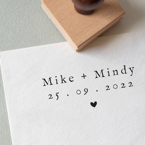 May include: A wooden stamp with a dark brown handle is positioned above a white paper. The paper displays the names "Mike + Mindy", the date "25.09.2022", and a small black heart. The stamp is partially visible.