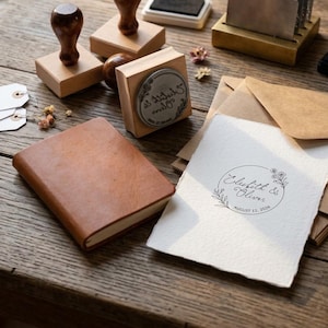 May include: A tan leather-bound notebook, several wooden rubber stamps, and a piece of textured white paper featuring a circular floral design with the text "Elizabeth & Oliver" and "August 12, 2026" printed in black ink, alongside brown envelopes.