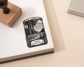 May include: A black and white Ex Libris bookplate sticker featuring a forest scene with a fox, two figures, and the text "JOHN WHITAKER". The sticker is on a white book, with a wooden stamp and a gray book in the background.