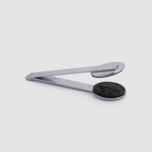 May include: A silver-colored bottle opener with a black circular base. The opener has a lever design with a circular black base on one end and a flat, rectangular base on the other. The black base has a detailed design.
