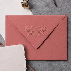 May include: A dusty rose coloured envelope with a deckled edge and a central embossed design featuring two floral stems flanking the letter M. The envelope is made of textured paper and is positioned alongside pieces of white deckled edge paper.