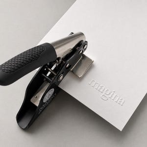 K&ouml;nnte beinhalten: Ein manuelles Pr&auml;gewerkzeug aus schwarzem und silbernem Metall mit einem strukturierten schwarzen Griff, das in ein dickes wei&szlig;es Kartenpapier dr&uuml;ckt. Das gepr&auml;gte Papier zeigt das Wort magina, das deutlich in einer Serifenschrift eingepr&auml;gt ist.