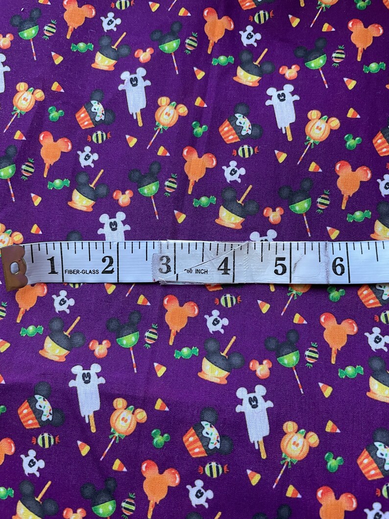 May include: Purple fabric with a repeating pattern of Mickey Mouse heads, candy corn, lollipops, and cupcakes. The fabric is measured with a tape measure that reads "60 inch".