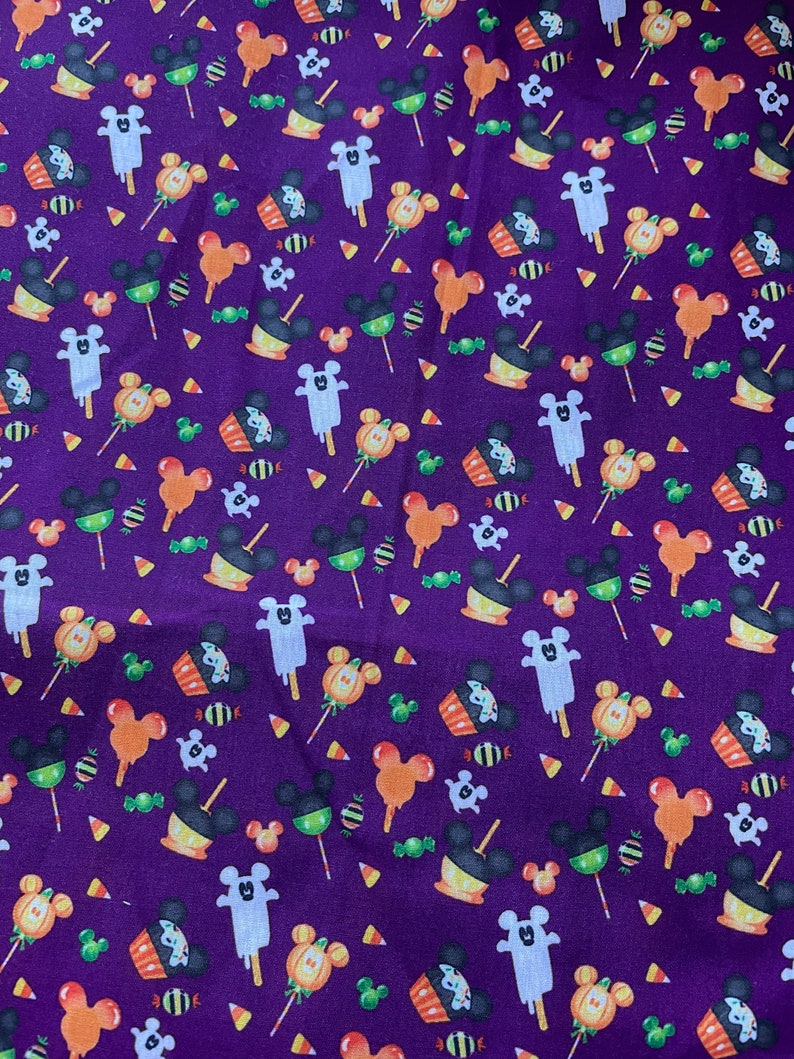 May include: Purple fabric with a repeating pattern of Mickey Mouse heads, ghosts, candy corn, cupcakes, and lollipops. The pattern is in shades of orange, yellow, green, white, and black.