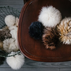 May include: Five fluffy pom poms in various colours, including white, black, brown, and beige, are arranged on a dark brown wooden surface.